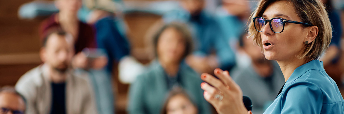 Woman wearing glasses, presenting to a room of people