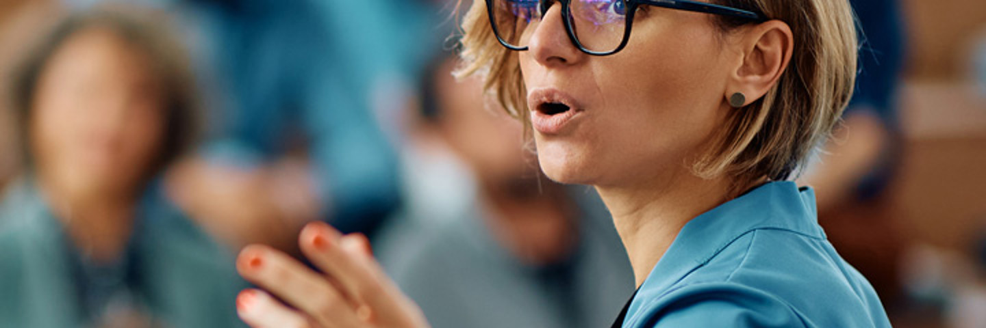 Woman wearing glasses, presenting to a room of people